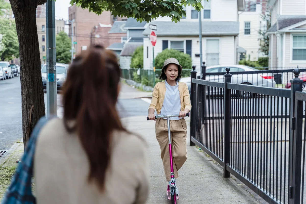 A young girl rides her scooter on a city sidewalk while her mother watches. The scene captures a fun outdoor moment.