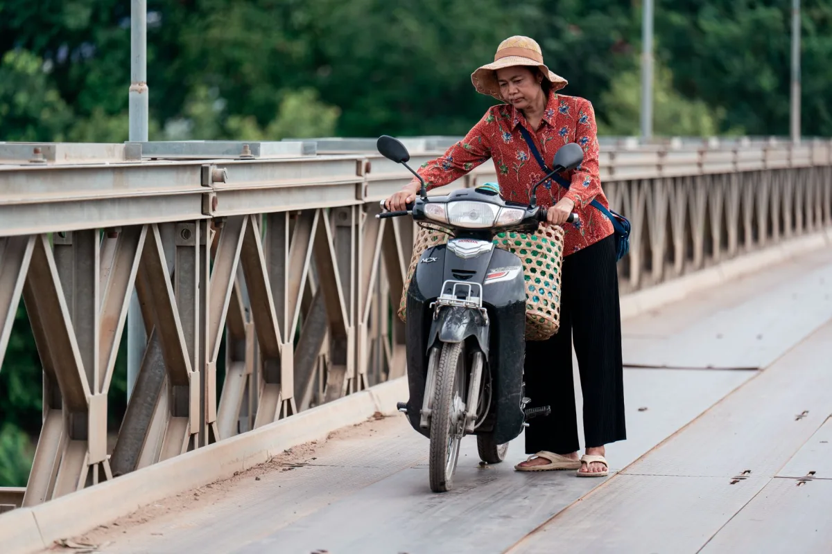 Elderly woman pushing her scooter on a metal bridge surrounded by greenery.